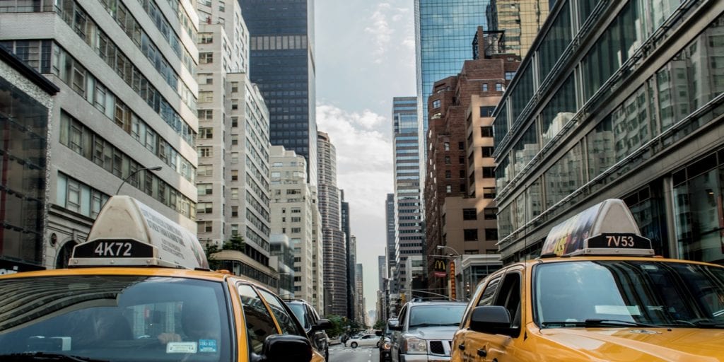A bustling city street lined with tall buildings features several yellow taxis in the foreground, navigating through traffic. The high-rise buildings reflect the urban environment, while the cloudy sky overhead adds to the metropolitan ambiance.