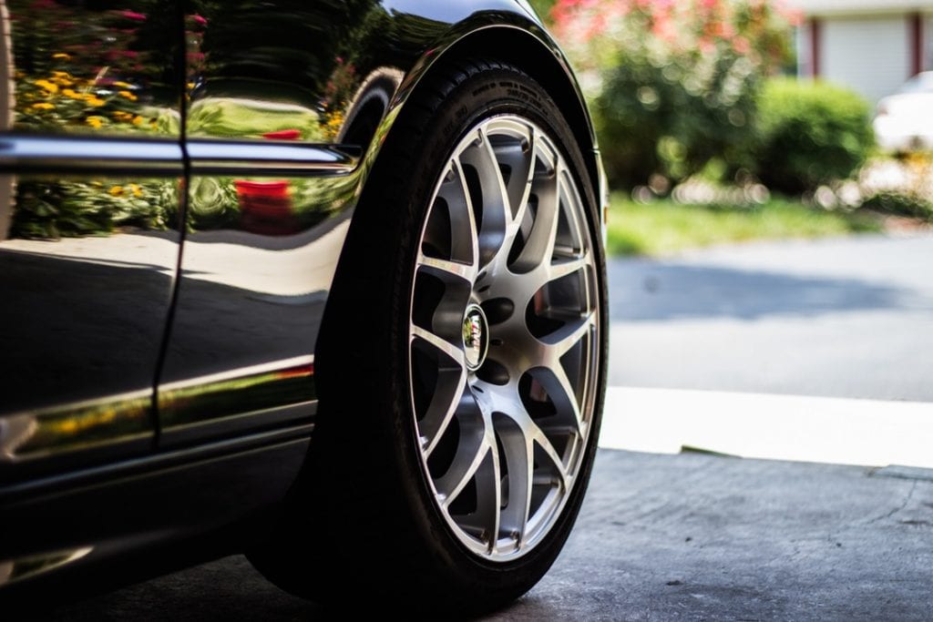 Close-up of a polished silver alloy wheel on a luxury car, highlighting its sleek design and reflective surface. The shiny black exterior of the car mirrors a vibrant garden with colorful flowers in the background, emphasizing the car's pristine condition. 