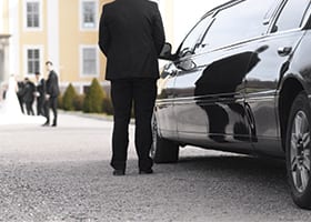 A sleek black limousine is parked outside a luxurious wedding venue. A chauffeur in a black suit stands beside the limo, awaiting the newlyweds. In the background, elegantly dressed wedding guests are seen, adding to the celebratory atmosphere. 